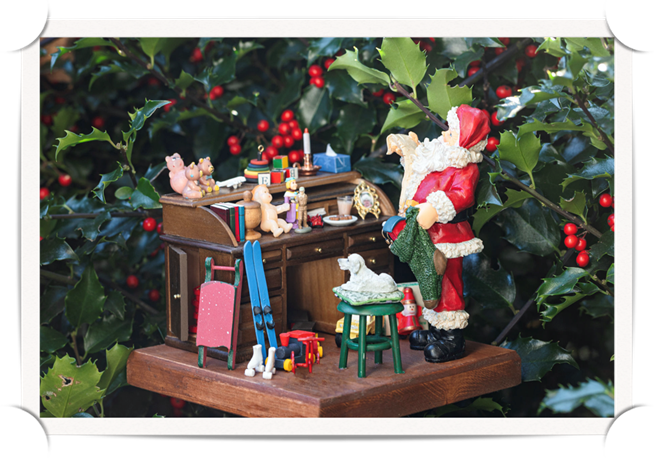 A miniature display of Father Christmas in his office preparing for delivery of gifts - all created by Freedom Miniatures, and displayed next to a holly bush with red berries